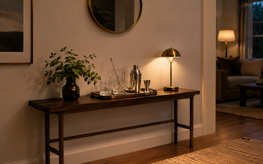 Wide establishing shot of a small home bar setup on a console shelf in evening light, warm palette of amber, brass and dark…