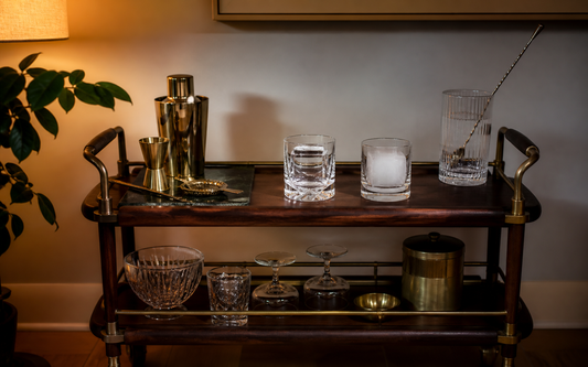 Wide establishing shot of a home bar cart in evening light, warm palette of amber, brass and dark wood against an off-white…