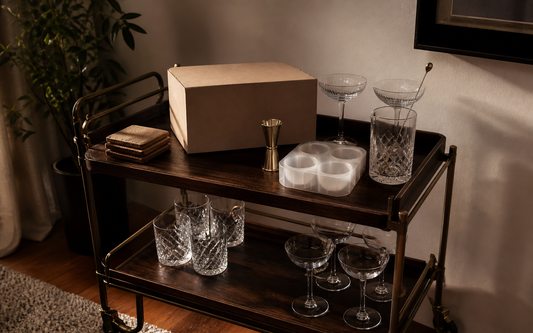 Wide establishing shot of a home bar cart in evening light, warm palette of amber, brass and dark wood against an off-white…