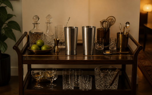 A home bar cart in evening light with two stainless Boston shaker tins standing side by side among real glassware and tools…