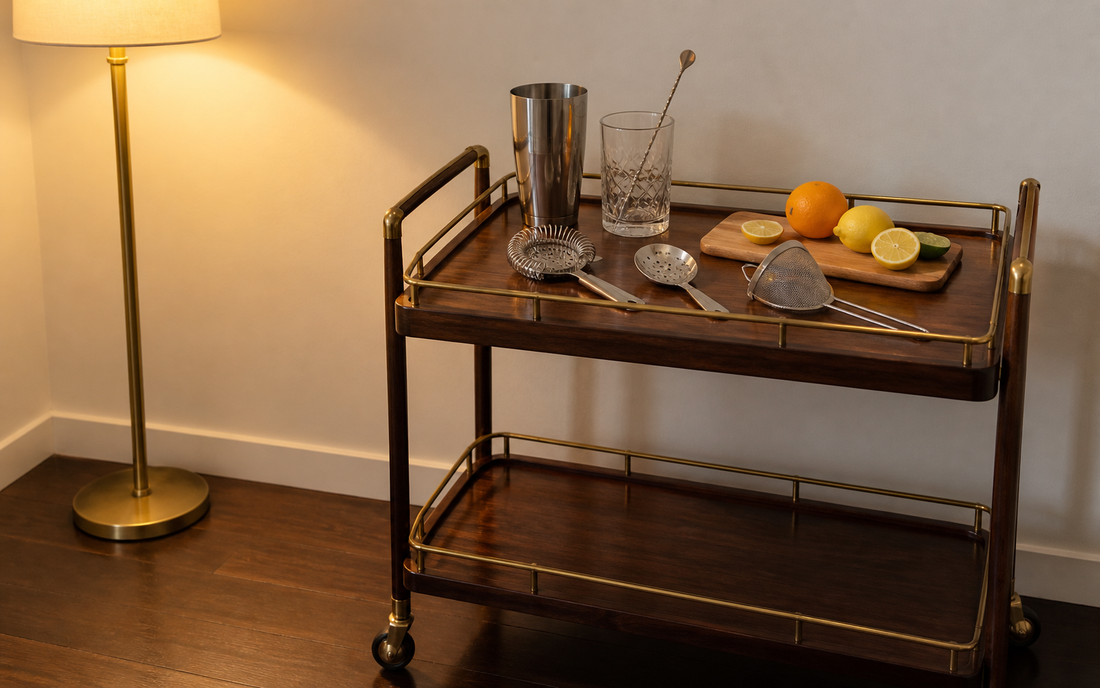 Wide establishing shot of a home bar cart in evening light, warm palette of amber, brass and dark wood against an off-white…