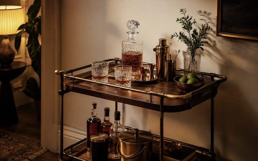Wide establishing shot of a home bar cart in evening light, a glass decanter with a fitted stopper standing beside two…