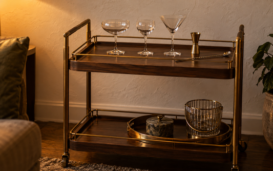 A small home bar cart with a dark walnut frame and brass rails against an off-white plaster wall, evening light from a warm…