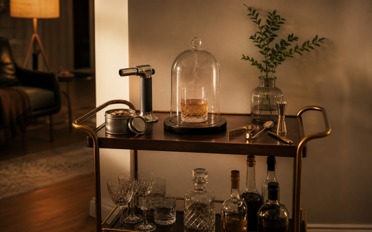 A home bar cart in evening light with a cocktail smoker kit laid out on top: a small canister of wood chips, a butane torch…