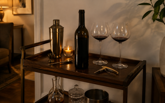 Wide establishing shot of a home bar cart in evening light, warm palette of amber, brass and dark wood against an off-white…