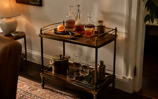 Wide establishing shot of a home bar cart in evening light, warm palette of amber, brass and dark wood against an off-white…