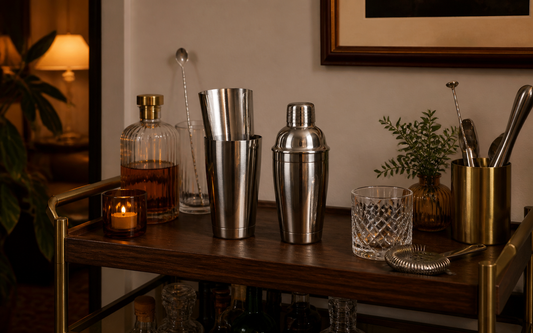 Wide establishing shot of a home bar cart in evening light, a two-piece Boston shaker (metal tins) and a three-piece cobbler…