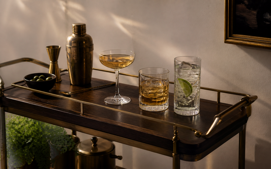 Wide establishing shot of a home bar cart in evening light, three types of glassware grouped together — a coupe, a rocks…