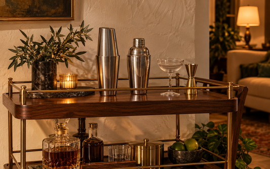 A home bar cart in evening light: dark walnut wood with brass rails, a stainless steel Boston shaker pair and a stainless…