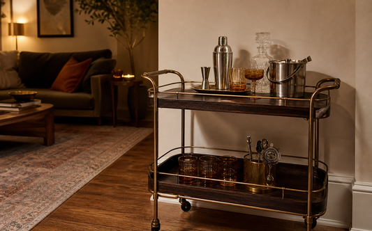 Wide establishing shot of a brass and dark wood bar cart in the corner of a warm-lit living room, holding a stainless boston…